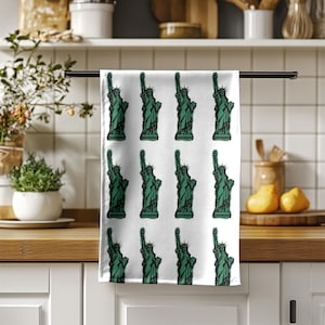 May include: White kitchen towel with a repeating pattern of the Statue of Liberty in green. The towel is hanging on a black bar in a kitchen setting. The Statue of Liberty is a famous American landmark.