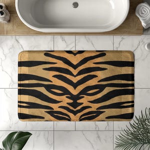 May include: Rectangular bath mat with a tiger stripe pattern. The mat features a tan background with black stripes, creating a bold and eye-catching design. The mat is designed to be placed in a bathroom setting.