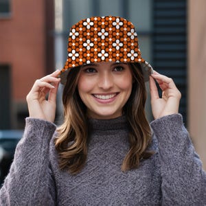 Könnte beinhalten: Ein Bucket Hat mit einem braunen Hintergrund und einem sich wiederholenden Muster aus orangefarbenen und weißen Blumenformen. Die Person trägt einen grauen Pullover und hält den Hut.
