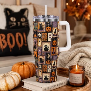 May include: A stainless steel tumbler with a handle and straw, featuring a repeating pattern of Halloween-themed rectangular tiles including black cats, jack-o'-lanterns, ghosts, skulls, spiderwebs, witch hats, and potion bottles on a dark background.