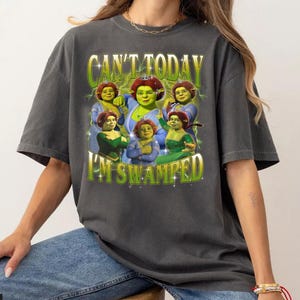 Puede incluir: Camiseta gris oscuro con un gráfico del personaje Fiona de la película animada Shrek. El texto "CAN'T TODAY I'M SWAMPED" está impreso en verde y dorado. El diseño incluye múltiples imágenes de Fiona.