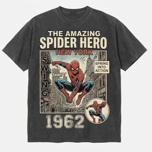 Pode incluir: T-shirt cinzenta escura com um gr&aacute;fico em estilo de banda desenhada vintage do Homem-Aranha. O design inclui o texto "THE AMAZING SPIDER HERO NEW YORK 1962" e "SPRING INTO ACTION". Uma imagem menor do Homem-Aranha est&aacute; no canto inferior direito.