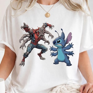 May include: White t-shirt with a graphic of a zombie-like Spider-Man and a blue alien character. The Spider-Man figure is in red, blue, and grey, while the alien is blue. The design is centered on the shirt.