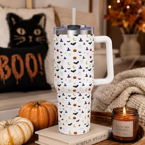Puede incluir: Un vaso térmico blanco de acero inoxidable con asa y pajita, que presenta un patrón repetitivo de calabazas naranjas, sombreros de bruja morados, murciélagos negros, lunas crecientes amarillas y fantasmas blancos. La tapa es transparente.