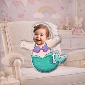 May include: A mermaid-themed pillow featuring a smiling baby with outstretched arms. The pillow has a teal tail and a purple seashell top. The baby is set against a nursery backdrop with a rainbow and a white armchair.