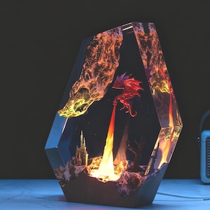 Może przedstawiać: Dekoracyjna lampa z żywicy o tematyce fantasy, przedstawiająca czerwonego smoka ziejącego ogniem. Lampa ma geometryczny kształt z drewnianą podstawą. Wnętrze jest oświetlone ciepłymi pomarańczowymi i żółtymi diodami LED, tworząc dramatyczny efekt. Mierzy około 20 cm wysokości.