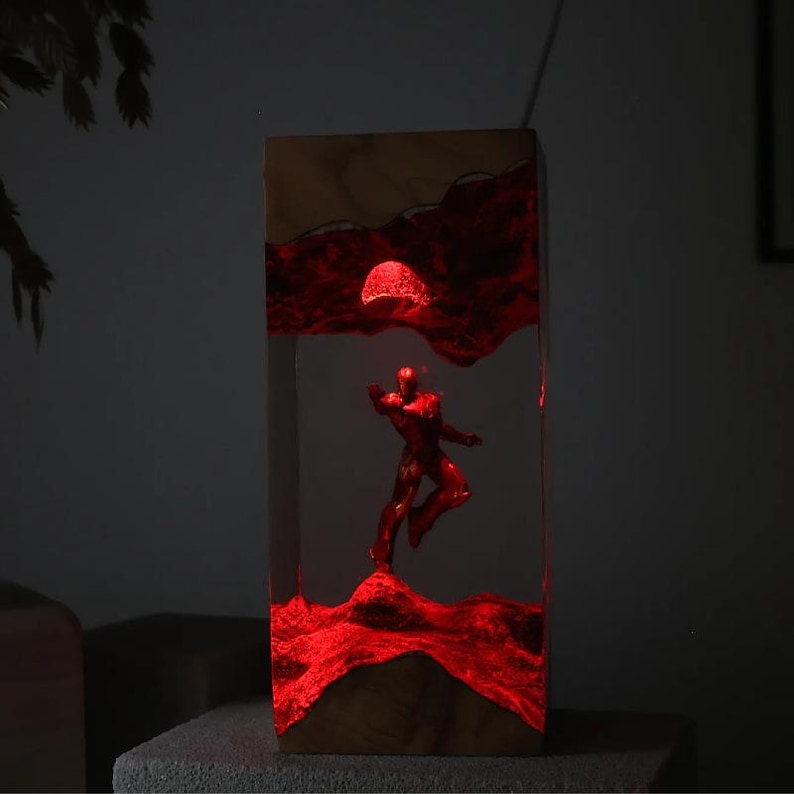 Puede incluir: Una l&aacute;mpara rectangular de madera con interior iluminado en rojo. Una figura roja est&aacute; colocada en el centro, con un paisaje iluminado en rojo y una esfera iluminada en rojo encima. La l&aacute;mpara est&aacute; sobre una superficie gris.