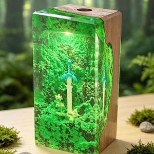 May include: A rectangular lamp featuring a block of translucent green resin embedded with a light-colored sword and a small plant, set against a natural wood base. The resin has a textured, moss-like appearance and is illuminated from within.