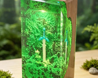 Könnte beinhalten: Eine rechteckige Lampe mit einem Block aus durchscheinendem grünem Harz, in den ein helles Schwert und eine kleine Pflanze eingebettet sind, auf einem Sockel aus Naturholz. Das Harz hat eine moosartige Textur und wird von innen beleuchtet.