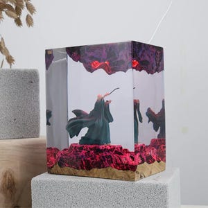 Può includere: Lampada a cubo in resina trasparente con una statuina di mago con una bacchetta. La lampada ha un design a formazione rocciosa rossa e viola nella parte superiore e inferiore, con una base in legno. Il mago indossa una veste verde.