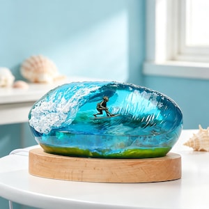 May include: A decorative resin sculpture featuring a miniature bronze-toned surfer riding a translucent blue ocean wave with white foam crests. The piece is mounted on a smooth, oval-shaped light-colored wooden base with a visible power cord.