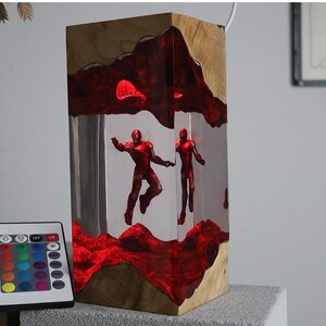 Puede incluir: L&aacute;mpara rectangular de madera con un centro de resina transparente, con dos figuras rojas de Iron Man. La resina est&aacute; iluminada con luz roja, y la l&aacute;mpara incluye un mando a distancia para ajustar el color. La base de madera tiene vetas naturales.