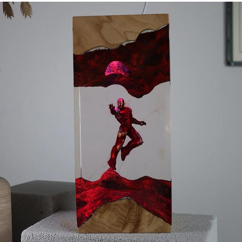 Puede incluir: Una pieza de arte rectangular de madera con una figura roja de Iron Man. La obra de arte incorpora resina roja y un marco de madera. El dise&ntilde;o incluye un elemento rojo brillante, posiblemente un sol o una fuente de luz, que a&ntilde;ade profundidad e inter&eacute;s visual a la pieza.