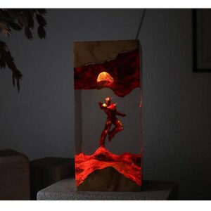 Puede incluir: L&aacute;mpara rectangular de madera y resina con una figura roja en pose din&aacute;mica, sobre un fondo rojo y naranja ardiente con un sol brillante. La l&aacute;mpara se ilumina desde dentro, creando un brillo ambiental c&aacute;lido.