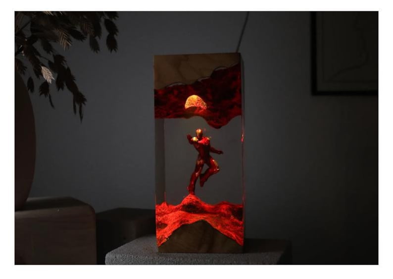 Puede incluir: L&aacute;mpara rectangular de madera y resina con una figura roja en pose din&aacute;mica, sobre un fondo rojo y naranja ardiente con un sol brillante. La l&aacute;mpara se ilumina desde dentro, creando un brillo ambiental c&aacute;lido.