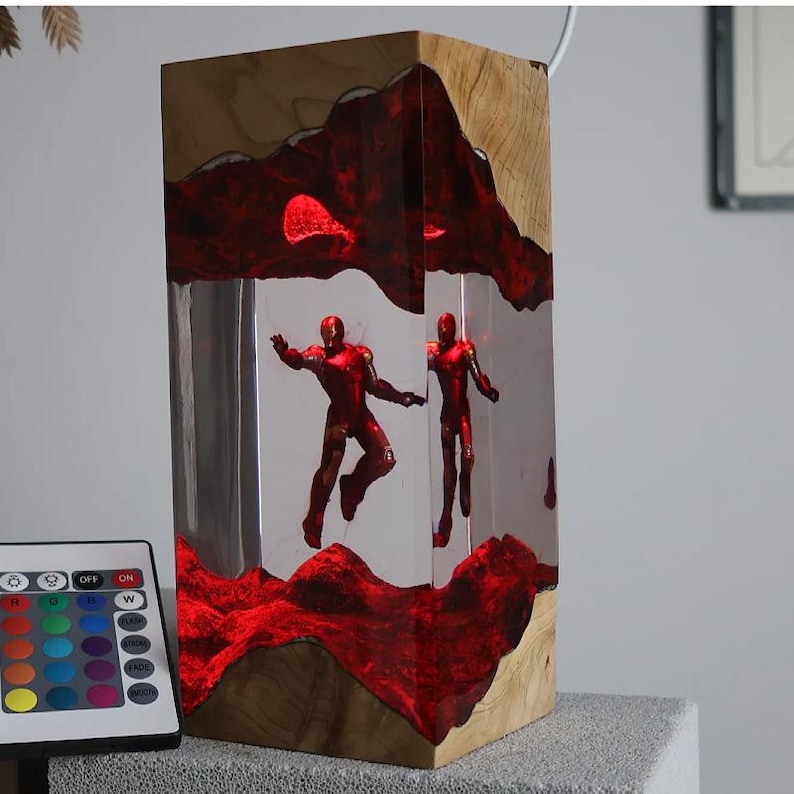 Puede incluir: L&aacute;mpara rectangular de madera con un centro de resina transparente, con dos figuras rojas de Iron Man. La resina est&aacute; iluminada con luz roja, y la l&aacute;mpara incluye un mando a distancia para ajustar el color. La base de madera tiene vetas naturales.