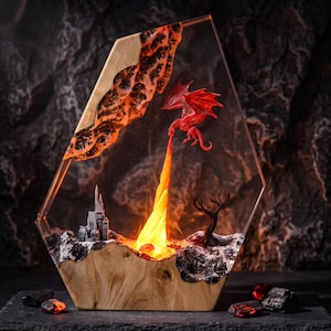 May include: A hexagonal resin and wood lamp featuring a red dragon figurine breathing a bright orange flame. The scene includes a miniature gray castle, a dark leafless tree, and textured rocky terrain embedded within the clear resin structure.