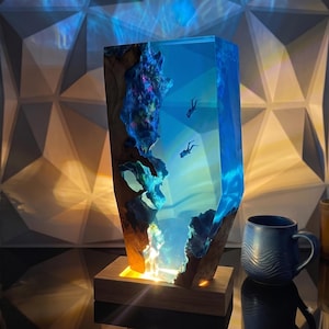 Könnte beinhalten: Eine rechteckige Lampe mit einem vertikalen Stück Naturholz, das in durchscheinendes blaues Harz eingegossen ist. Das Design zeigt eine Unterwasserszene mit zwei kleinen Taucherfiguren auf einem beleuchteten Holzsockel.