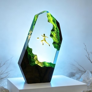 May include: A vertical, multifaceted resin and wood sculpture featuring a suspended, golden-colored figure in a dynamic soccer pose. The piece incorporates translucent, vibrant green resin sections that contrast with dark, textured wood elements.