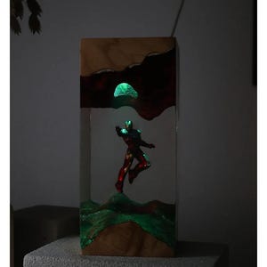 Puede incluir: Bloque de madera con una figura de Iron Man brillante. La obra de arte incorpora elementos brillantes verdes y rojos, con un paisaje texturizado y un orbe verde brillante. La pieza est&aacute; sobre un fondo neutro.