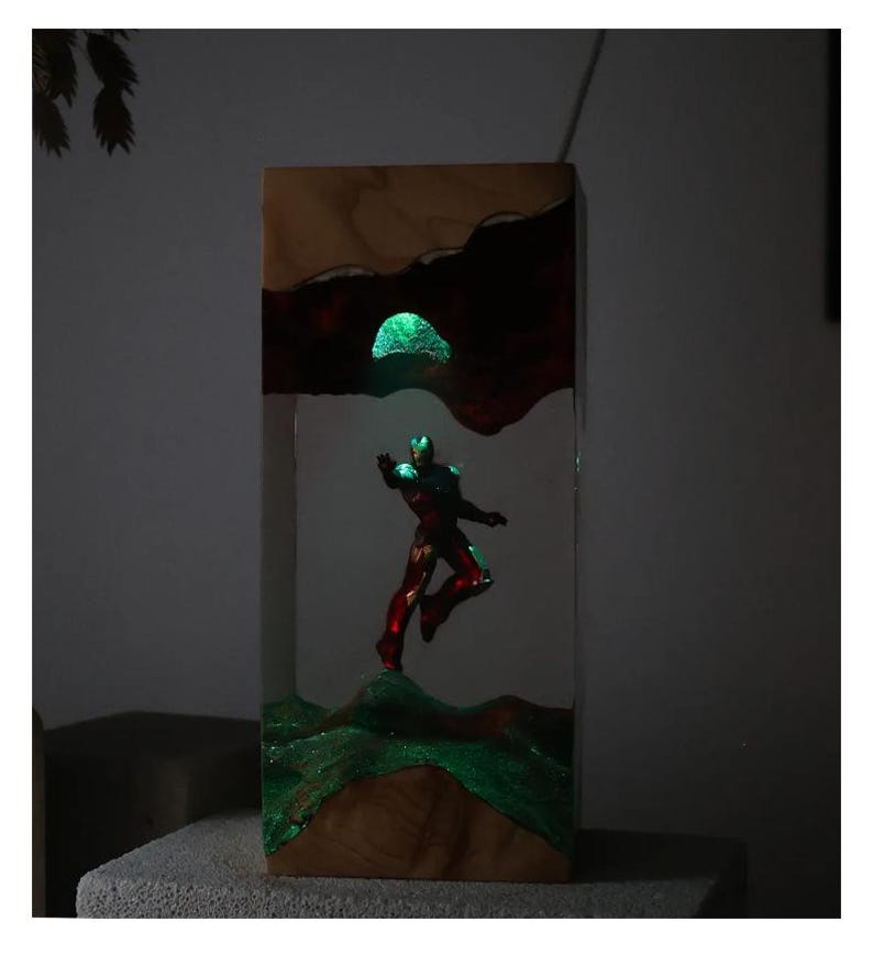 Puede incluir: Bloque de madera con una figura de Iron Man brillante. La obra de arte incorpora elementos brillantes verdes y rojos, con un paisaje texturizado y un orbe verde brillante. La pieza est&aacute; sobre un fondo neutro.