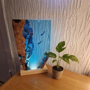Könnte beinhalten: Dekorative Holzlampe mit blauem Harzdesign, das zwei Tauchersilhouetten zeigt. Die Lampe hat einen Holzsockel und steht auf einem Holztisch. Eine kleine Topfpflanze befindet sich neben der Lampe.