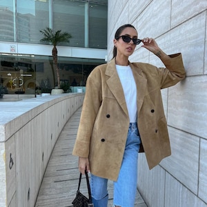 May include: A tan suede double-breasted blazer with black buttons, worn over a white t-shirt and light blue distressed denim jeans. The outfit is paired with black cat-eye sunglasses and a small black patterned shoulder bag with a silver buckle.