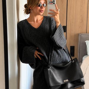 Puede incluir: Un suéter de punto acanalado gris carbón con cuello en V y mangas anchas, combinado con pantalones de punto acanalado a juego. Un bolso de hombro grande de cuero negro con un cierre giratorio rectangular plateado y correas finas.