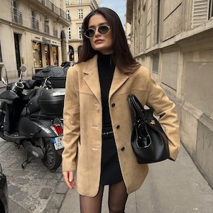 May include: A woman wearing a tan suede jacket, a black turtleneck sweater, a black mini skirt, black tights, and black boots. She is carrying a black leather handbag and wearing oval-shaped sunglasses.