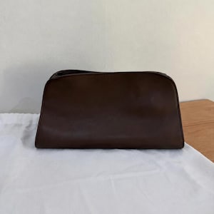 Op de afbeelding: Een donkerbruine rechthoekige tas met een gladde, matte afwerking. De tas heeft een gestructureerde bovenrand met een ritssluiting, een licht taps toelopende onderkant en strakke, minimalistische lijnen in het gehele ontwerp.