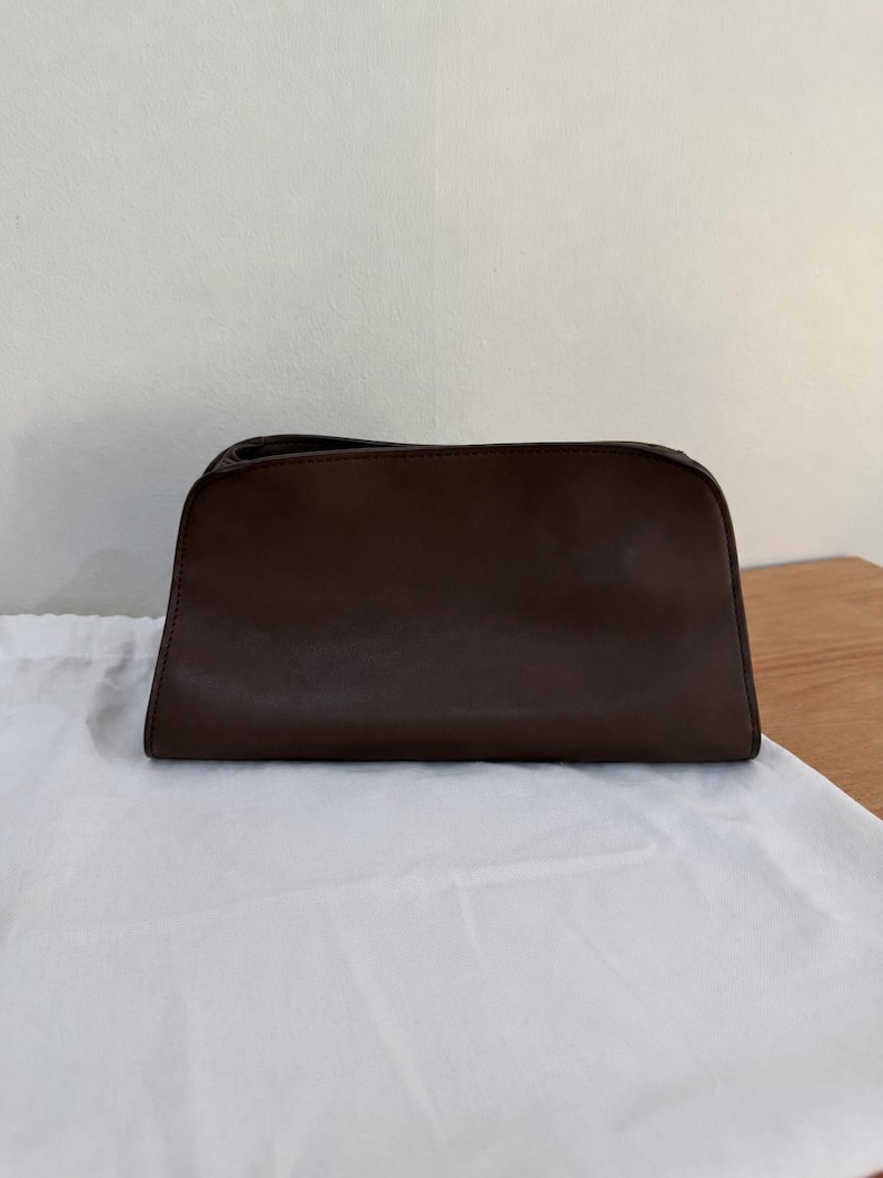 Op de afbeelding: Een donkerbruine rechthoekige tas met een gladde, matte afwerking. De tas heeft een gestructureerde bovenrand met een ritssluiting, een licht taps toelopende onderkant en strakke, minimalistische lijnen in het gehele ontwerp.