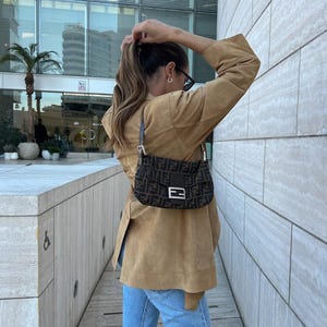 May include: A brown suede jacket with a tan hue, paired with light blue denim jeans and a dark brown shoulder bag featuring a repeating black logo pattern, a central rectangular silver-toned metal buckle, and a short adjustable leather strap.