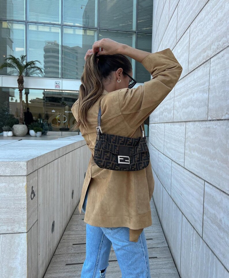 May include: A brown suede jacket with a tan hue, paired with light blue denim jeans and a dark brown shoulder bag featuring a repeating black logo pattern, a central rectangular silver-toned metal buckle, and a short adjustable leather strap.