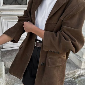May include: A brown suede blazer with a button fastening and a single pocket on the left side. The blazer is worn over a white t-shirt and black shorts.