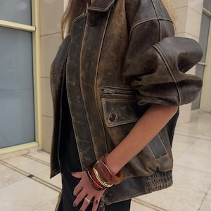 Pode incluir: Casaco de couro castanho com acabamento envelhecido. O casaco tem fecho de correr, um pequeno bolso e bainha canelada. As mangas são franzidas nos punhos. O casaco é usado sobre um vestido preto. A pessoa está usando várias pulseiras.