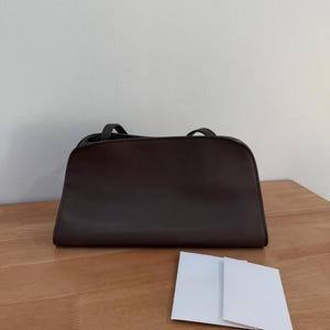 Op de afbeelding: Een donkerbruine rechthoekige handtas met een gladde afwerking, voorzien van een gestructureerd silhouet, een platte bodem en twee dunne schouderbanden aan de bovenkant. De tas staat horizontaal met een klein wit kaartje ervoor.