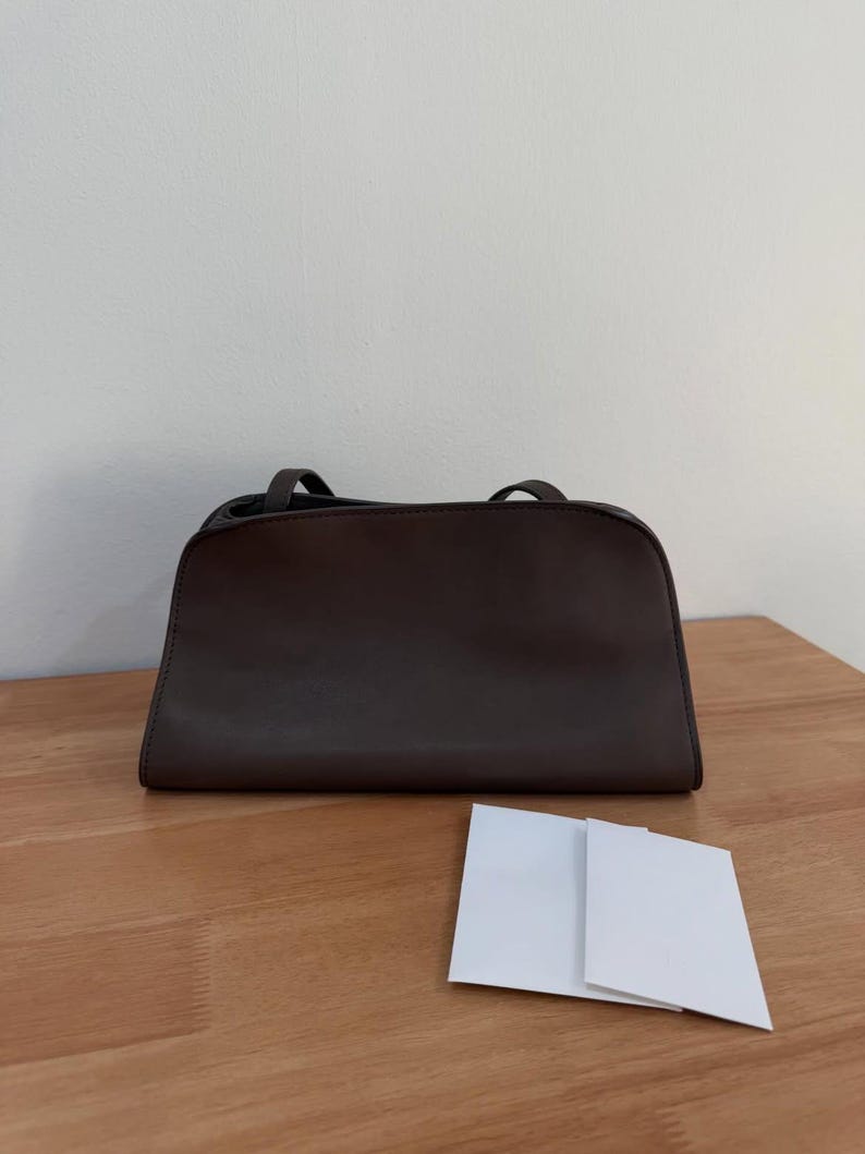 Op de afbeelding: Een donkerbruine rechthoekige handtas met een gladde afwerking, voorzien van een gestructureerd silhouet, een platte bodem en twee dunne schouderbanden aan de bovenkant. De tas staat horizontaal met een klein wit kaartje ervoor.