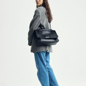 May include: A large black leather shoulder bag with a rectangular flap, a silver-toned metal clasp, and a thin shoulder strap. The bag is worn over a grey textured blazer, blue denim jeans, and black shoes, with thin-rimmed glasses.