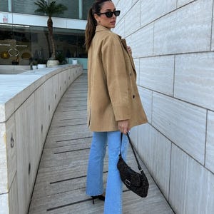 May include: A tan suede-textured jacket with a collar and buttons, paired with light blue denim jeans. A dark brown patterned shoulder bag with a silver buckle and black pointed-toe heels complete the outfit. The look includes black sunglasses.