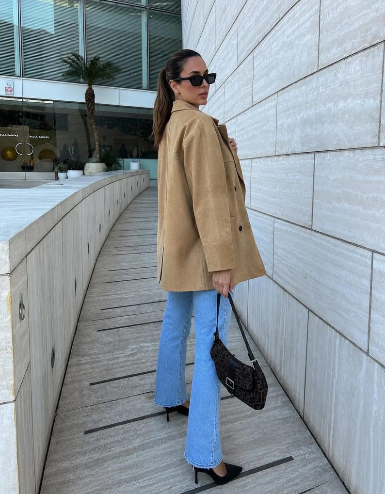 May include: A tan suede-textured jacket with a collar and buttons, paired with light blue denim jeans. A dark brown patterned shoulder bag with a silver buckle and black pointed-toe heels complete the outfit. The look includes black sunglasses.
