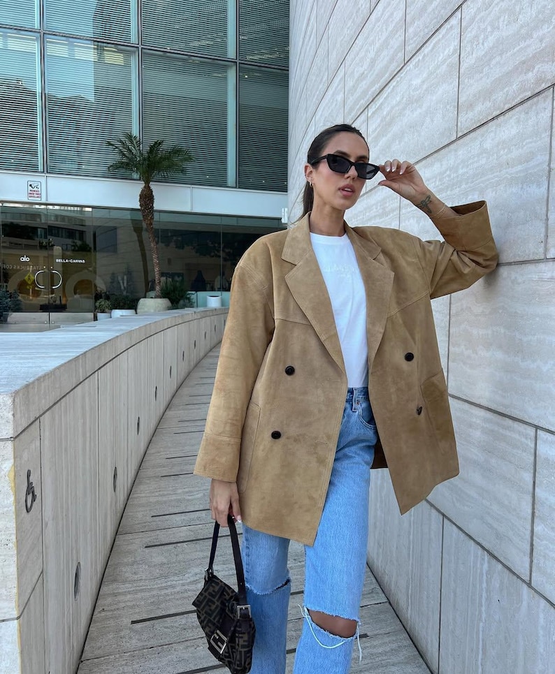 May include: A tan suede double-breasted blazer with black buttons, worn over a white t-shirt and light blue distressed denim jeans. The outfit is paired with black cat-eye sunglasses and a small black patterned shoulder bag with a silver buckle.