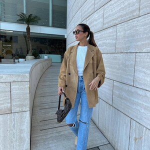 May include: A tan suede double-breasted blazer worn over a plain white t-shirt and light blue distressed denim jeans with knee rips. A dark brown patterned shoulder bag with a short strap is held in one hand, and black sunglasses are worn.