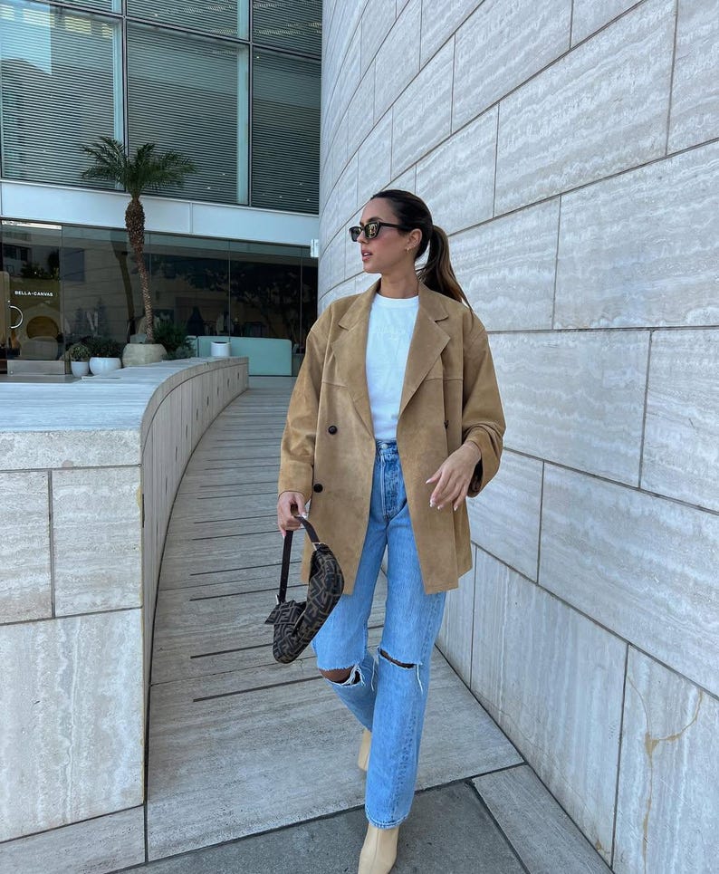May include: A tan suede double-breasted blazer worn over a plain white t-shirt and light blue distressed denim jeans with knee rips. A dark brown patterned shoulder bag with a short strap is held in one hand, and black sunglasses are worn.