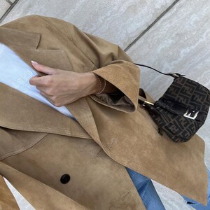 May include: A tan suede double-breasted blazer worn over a white t-shirt with blue jeans, paired with a small brown and black monogram shoulder bag featuring a silver buckle and a thin black strap, captured from a high angle.