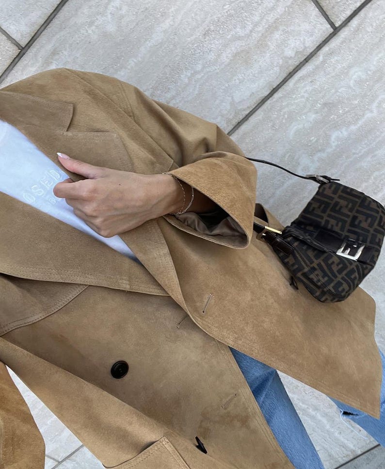 May include: A tan suede double-breasted blazer worn over a white t-shirt with blue jeans, paired with a small brown and black monogram shoulder bag featuring a silver buckle and a thin black strap, captured from a high angle.