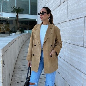 May include: A tan double-breasted suede blazer with notched lapels and four black buttons, worn over a plain white t-shirt and light blue distressed denim jeans with knee rips, paired with a small black shoulder bag and dark sunglasses.