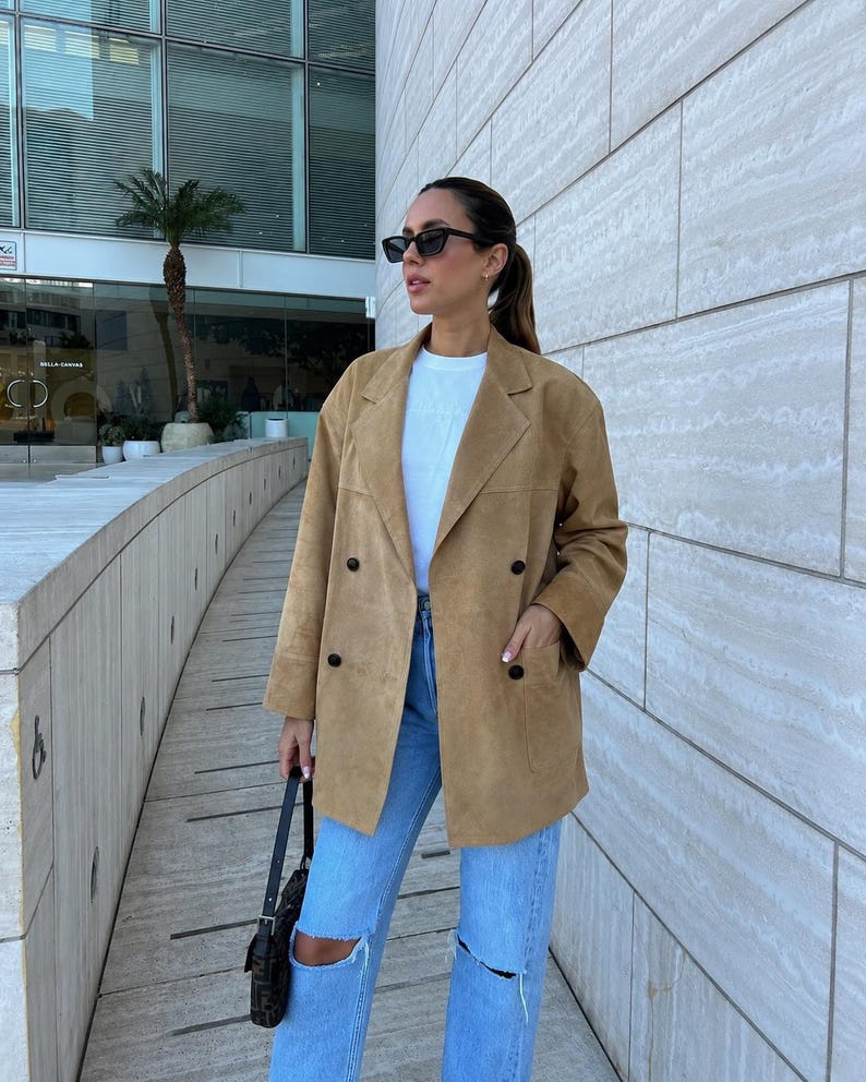 May include: A tan double-breasted suede blazer with notched lapels and four black buttons, worn over a plain white t-shirt and light blue distressed denim jeans with knee rips, paired with a small black shoulder bag and dark sunglasses.