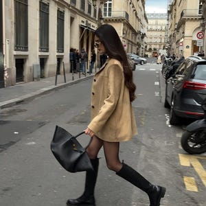 Pu&ograve; includere: Una donna con una giacca di camoscio marrone, collant neri e stivali neri al ginocchio cammina per una strada di citt&agrave; portando una borsa tote in pelle nera.