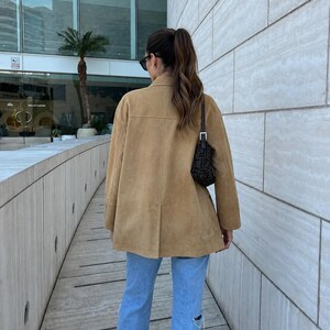 May include: A tan suede jacket with a collared neckline and long sleeves, paired with light blue denim jeans featuring distressed knee detailing. A small black shoulder bag with a repeating dark pattern is worn over the shoulder.