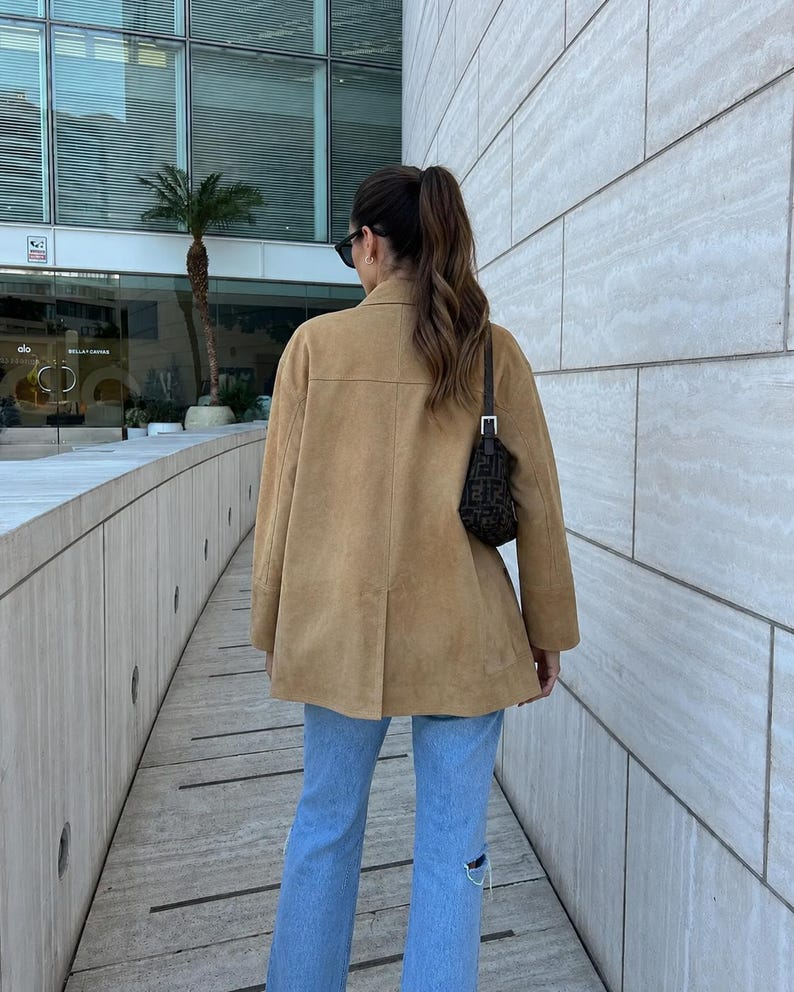 May include: A tan suede jacket with a collared neckline and long sleeves, paired with light blue denim jeans featuring distressed knee detailing. A small black shoulder bag with a repeating dark pattern is worn over the shoulder.